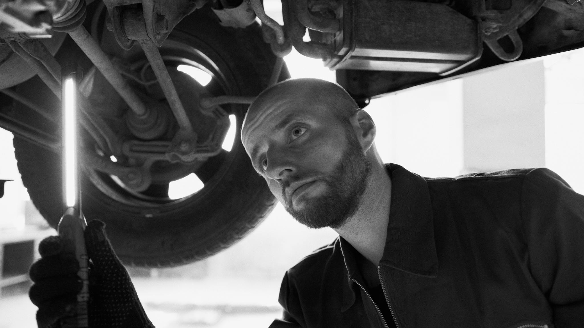 A mechanic carefully inspects under a vehicle in a workshop, utilizing a light in a grayscale setting.